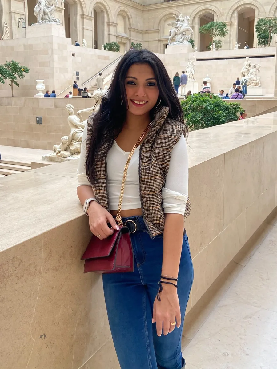 Shilpi Chakravorty leaning against a stone ledge in a sculpture court, in a houndstooth gilet and jeans.